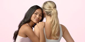 Zwei Junge Frauen mit langen Haaren vor rosa Hintergrund, eine Frau trägt ihr Haar offen, die andere Frau hat ihr Haar zum Zopf gebunden als Sinnbild für Tranktionsalopezie und Haarausfall durch Zopf und Dutt